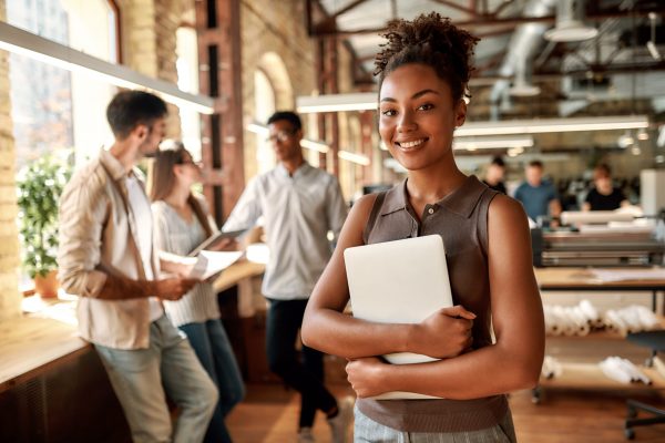 Happy to work here. Young and cheerful afro american woman holding laptop and smiling while standing in the modern office. Business concept. Job